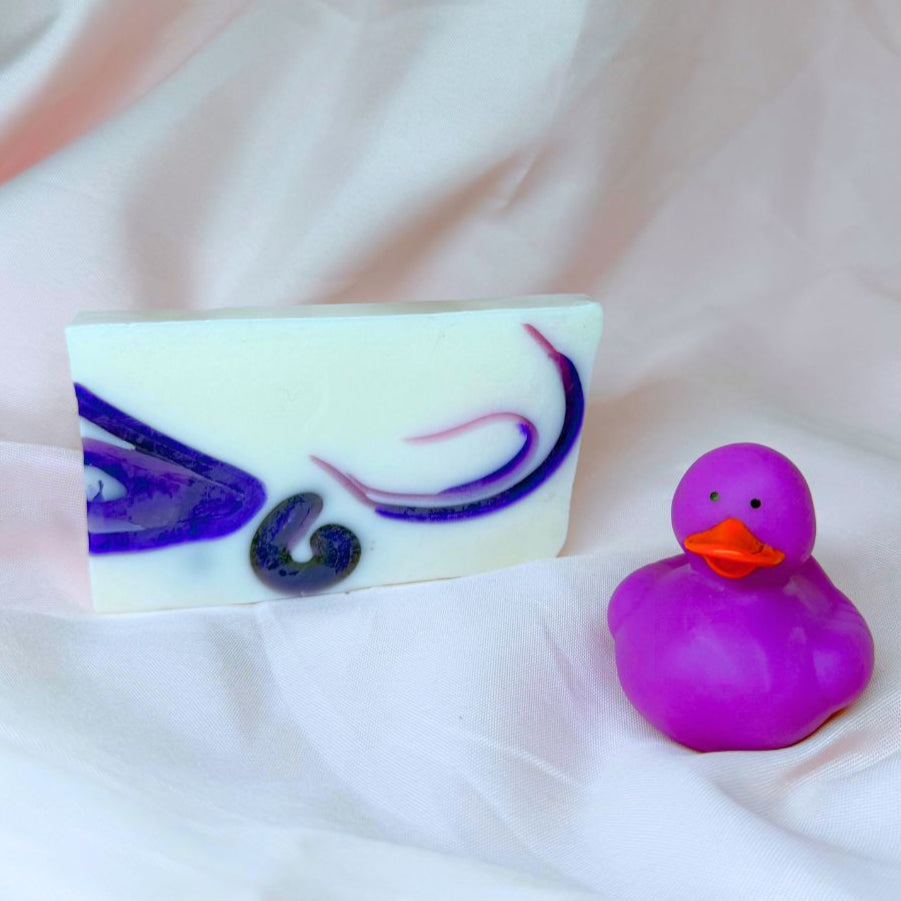 A bar of Primal Elements glycerin Ariel soap. It is white with swirls of purple. To the front right of the bar sits a purple rubber duck.