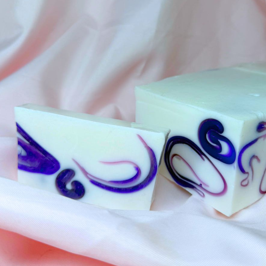 A bar of Primal Elements glycerin Ariel soap. It is white with swirls of purple. To the right of the bar is the loaf it was cut from, which bears a similar but not totally identical design.