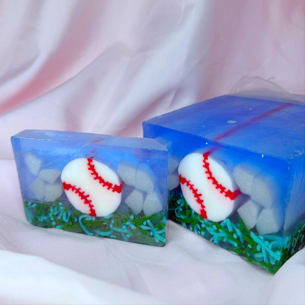 A bar of Primal Elements glycerin soap. The design is a baseball, white with red stitching, resting on some grass. The rest of the bar is blue and shite, like sky and clouds. To the right of the bar is the loaf it was cut from. It has the same design.