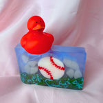A bar of Primal Elements glycerin soap. The design is a baseball, white with red stitching, resting on some grass. The rest of the bar is blue and shite, like sky and clouds. On top of the bar sits a red rubber duck.
