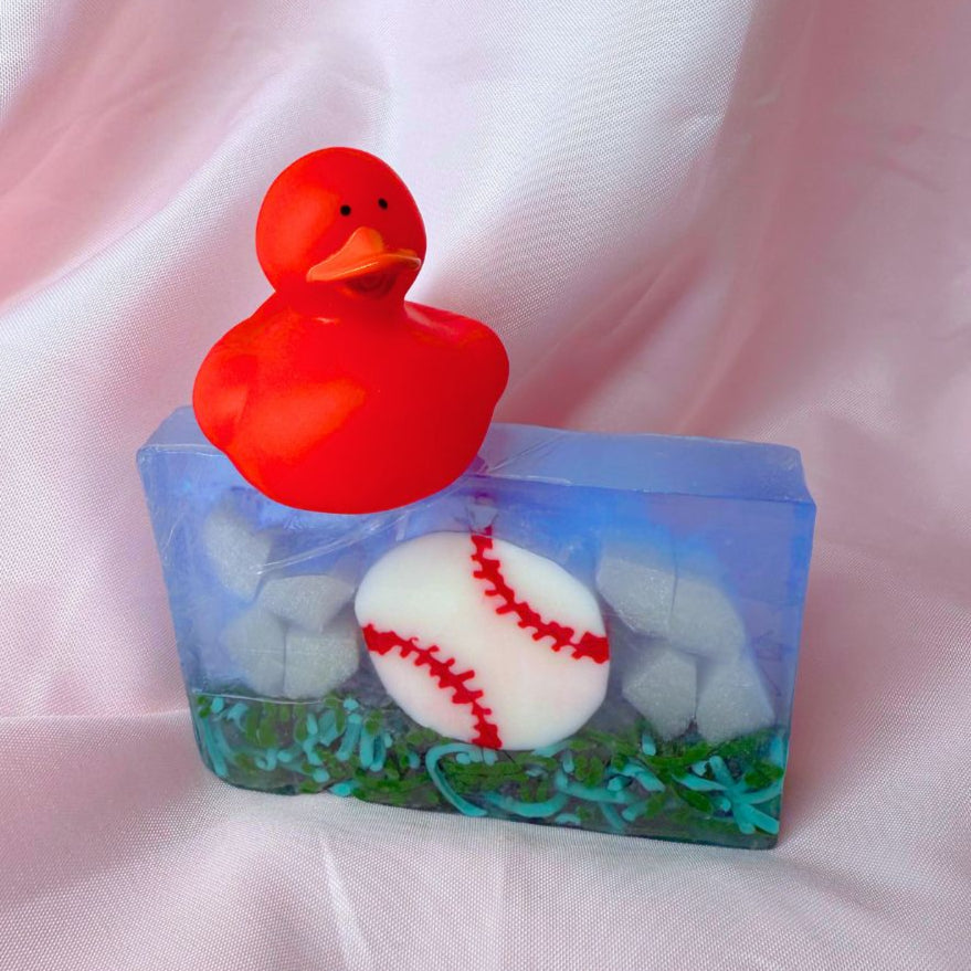 A bar of Primal Elements glycerin soap. The design is a baseball, white with red stitching, resting on some grass. The rest of the bar is blue and shite, like sky and clouds. On top of the bar sits a red rubber duck.