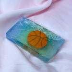A Primal Elements glycerin bar soap with the design of an orange basketball. The rest of the bar is blue with white pieces that look like the basketball net.