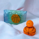Background: A Primal Elements glycerin bar soap with the design of an orange basketball. The rest of the bar is blue with white pieces that look like the basketball net. Foreground: a small rubber duck with a basketball design. It is orange with black lines and it is covered in small bumps.