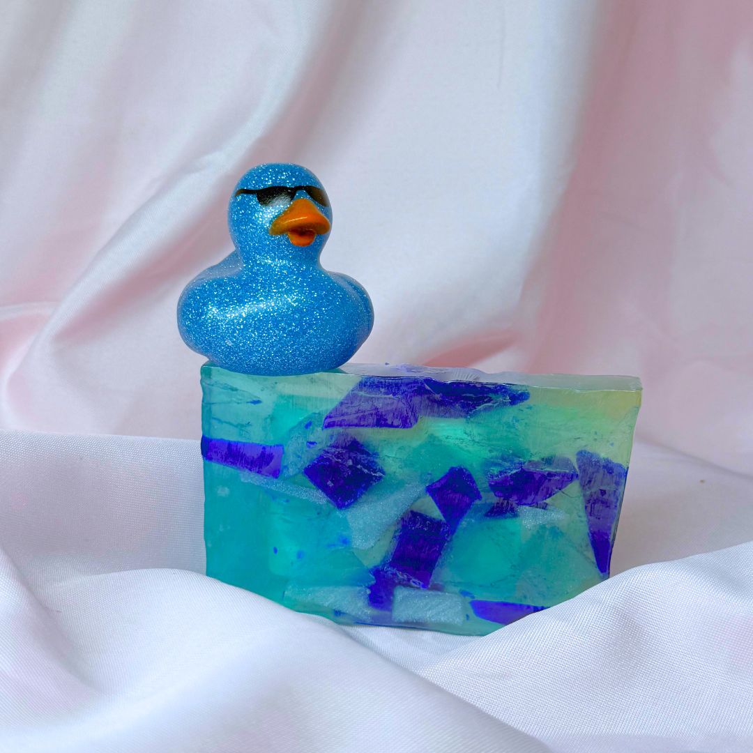 A bar of Primal Elements glycerin soap with the Beach Glass design. Various shapes and shades of blue decorate this bar, meant to look like sea glass in the ocean. On top of the bar sits a sparkly blue rubber duck wearing sunglasses.