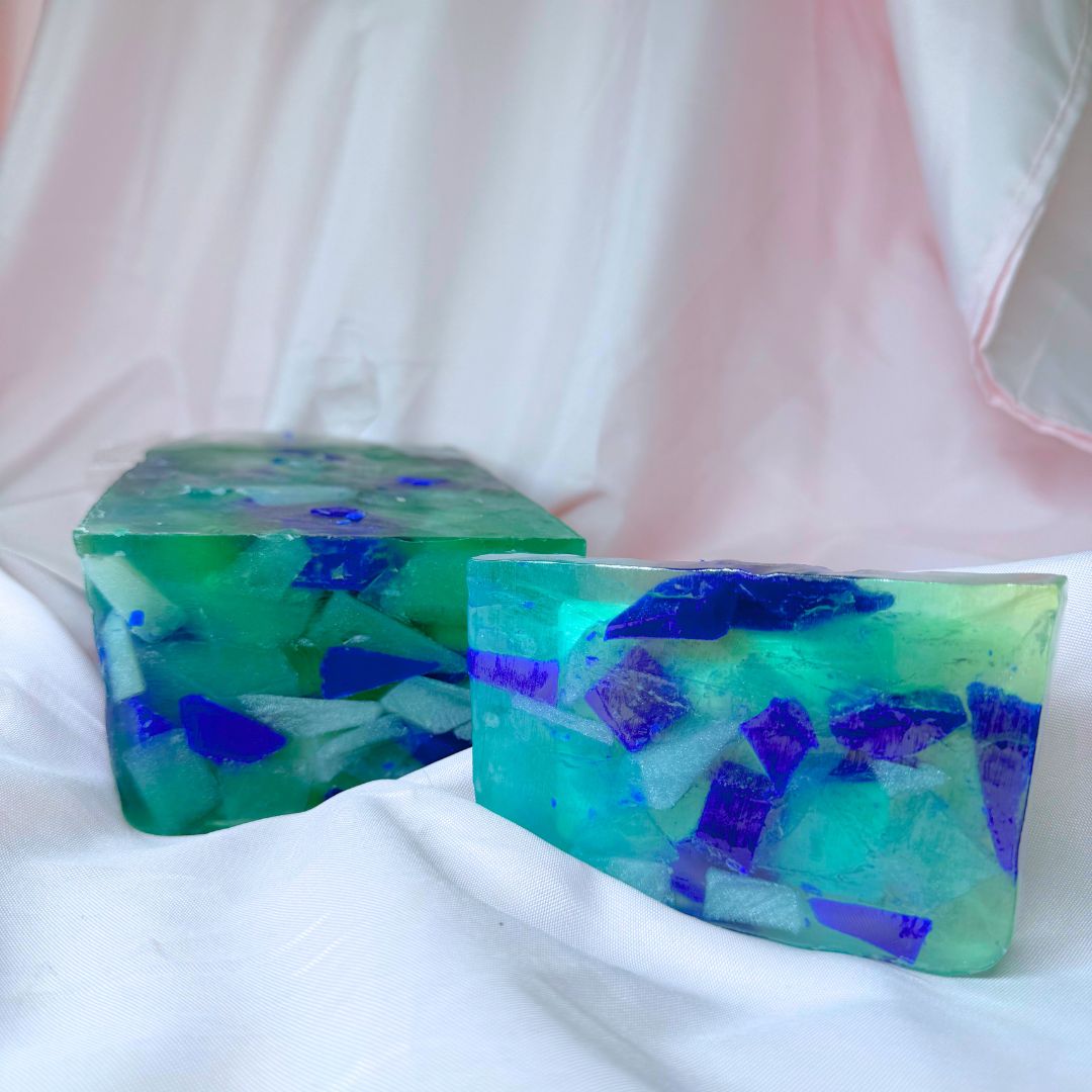 A bar of Primal Elements glycerin soap with the Beach Glass design. Various shapes and shades of blue decorate this bar, meant to look like sea glass in the ocean. To the bar's left is the loaf it was cut from, which bears the same design.