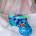 A bar of Primal Elements glycerin soap with the Beach Glass design. Various shapes and shades of blue decorate this bar, meant to look like sea glass in the ocean. In the front right of the bar is a sparkly blue rubber duck wearing sunglasses.