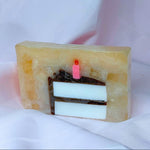 A bar of Primal Elements glycerin soap. Its design is a slice of birthday cake, with white cake, brown frosting, and a lit pink candle. The rest of the bar is a sparkly tan color.