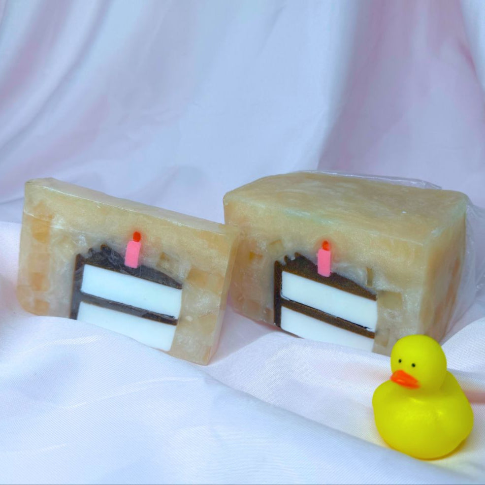 On the left, a bar of Primal Elements glycerin soap. Its design is a slice of birthday cake, with white cake, brown frosting, and a lit pink candle. The rest of the bar is a sparkly tan color. On the right, the loaf of soap that the bar was cut from. It has the same design. Even further right is a super mini rubber duck in traditional yellow.