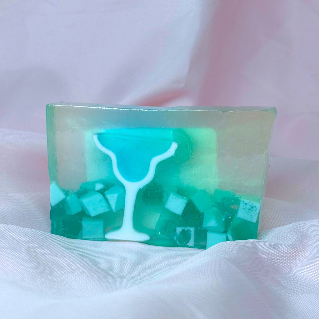 A bar of Primal Elements glycerin soap. Its design is of a margarita glass filled with blue liquid, with a lime slice on the rim. The rest of the bar is clear and translucent teal and there are some chunks of white floating at the bottom of the bar.