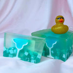 On the left, a bar of Primal Elements glycerin soap. Its design is of a margarita glass filled with blue liquid, with a lime slice on the rim. The rest of the bar is clear and translucent teal and there are some chunks of white floating at the bottom of the bar. On the right, the loaf that the bar came from, with a sparkly gold rubber duck in sunglasses resting on top of the loaf.