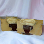 A bar of Primal Elements soap with a design of a steaming mug of coffee. Various shades of brown and visible coffee grounds. Behind it is the larger loaf that it was cut from, which has the same design.