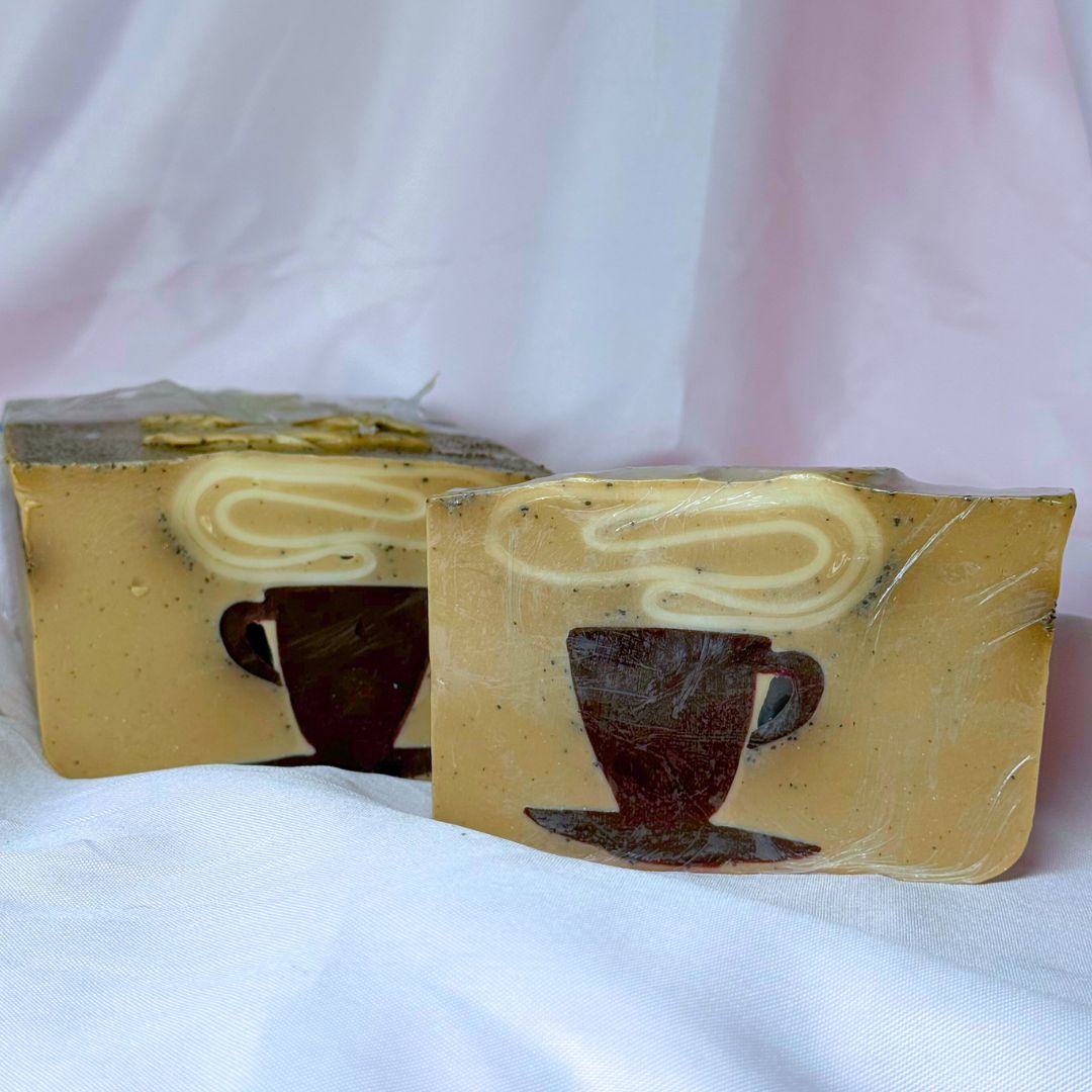 A bar of Primal Elements soap with a design of a steaming mug of coffee. Various shades of brown and visible coffee grounds. Behind it is the larger loaf that it was cut from, which has the same design.