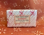 A rectangular bar of Michel Design Works Peppermint Shea Butter Soap wrapped in white paper with red diagonal stripes and candy cane motifs, placed on a rust-colored patterned fabric background.