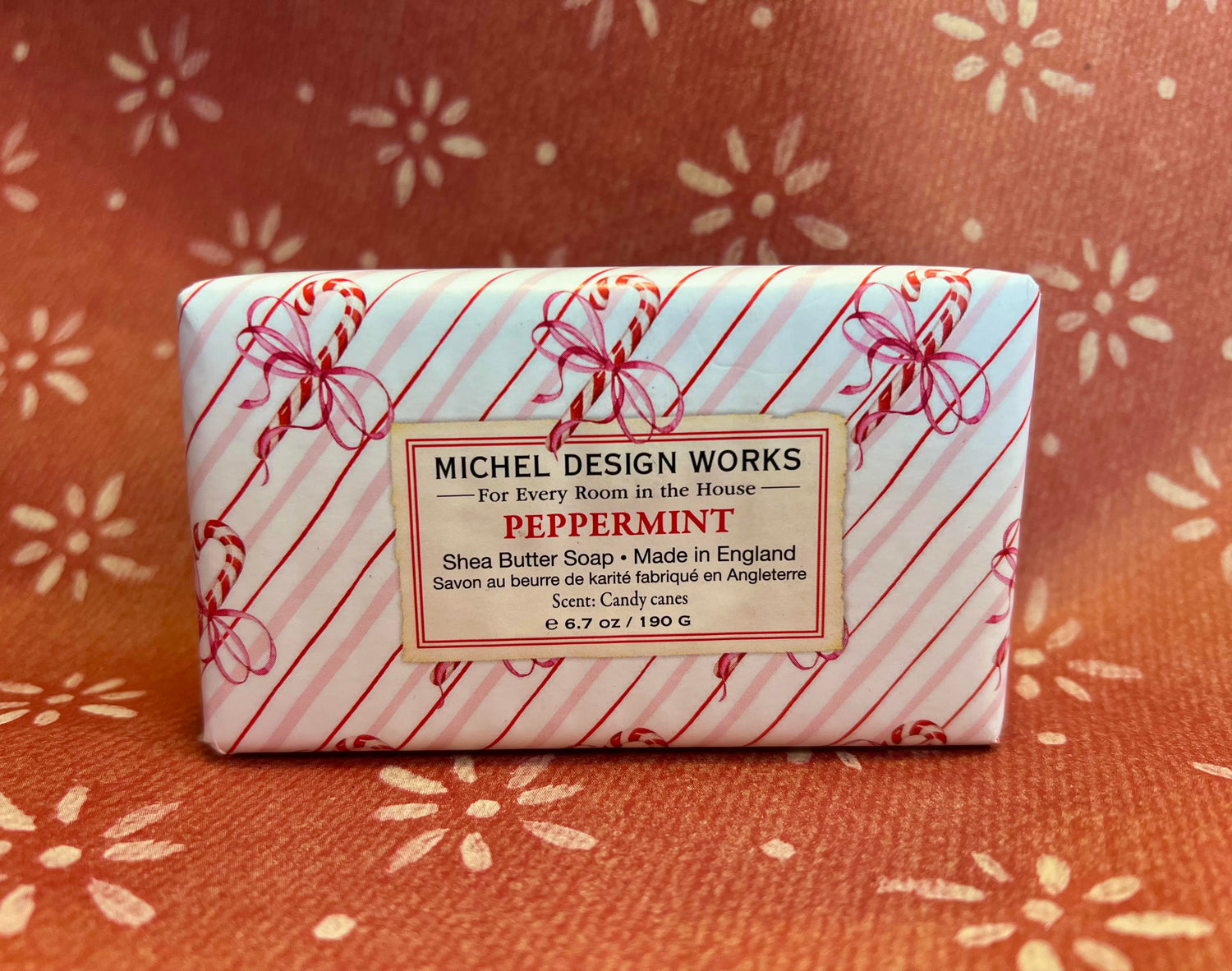 A rectangular bar of Michel Design Works Peppermint Shea Butter Soap wrapped in white paper with red diagonal stripes and candy cane motifs, placed on a rust-colored patterned fabric background.