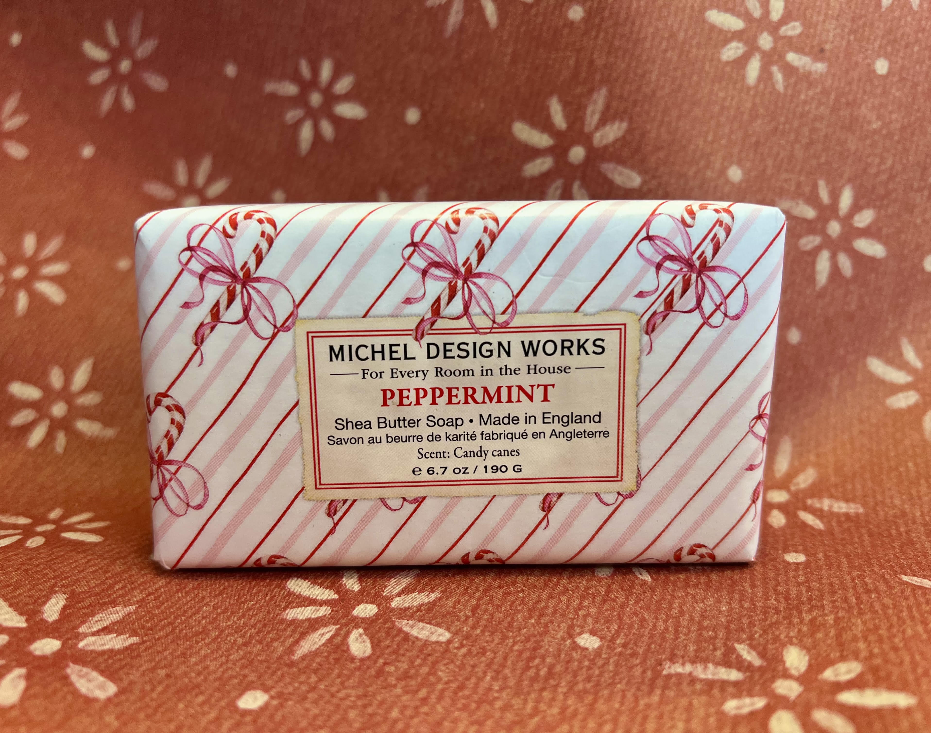 A rectangular bar of Michel Design Works Peppermint Shea Butter Soap wrapped in white paper with red diagonal stripes and candy cane motifs, placed on a rust-colored patterned fabric background.