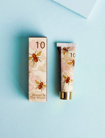 A cosmetic tube and matching rectangular box with a floral and bee print, labeled '10' and 'Honey & The Moon,' placed on a light blue background.
