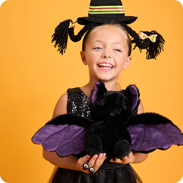 Child in a Halloween costume holding a black bat plush against an orange background. Bat is a heating pad and lavender scented