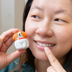 A close-up of a woman holding a dog-shaped lip balm near her face, pointing to her lips.