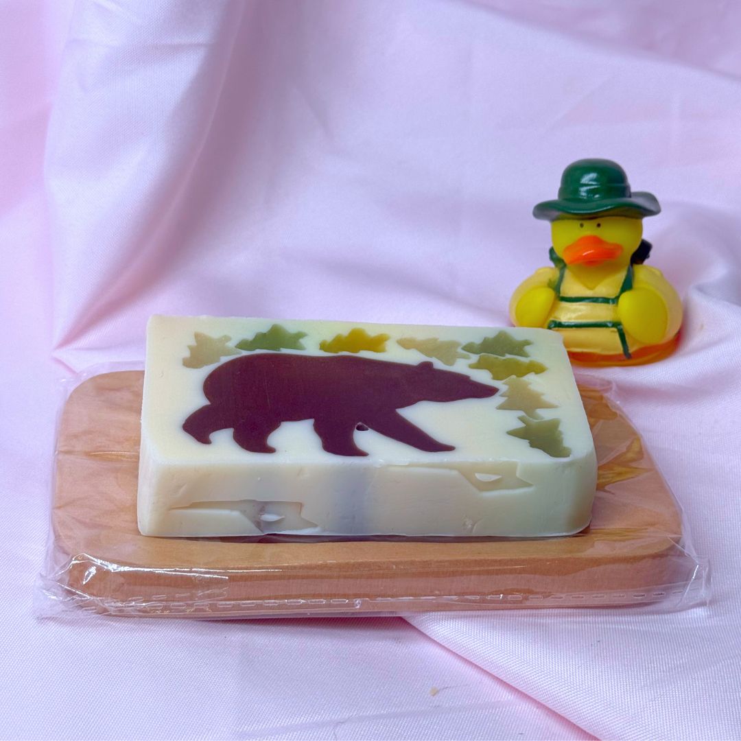 Bar of vegetable glycerin soap with a bear and tree design on a white background woodsy outdoors hiking camping summer on wooden soap dish with camping rubber ducky