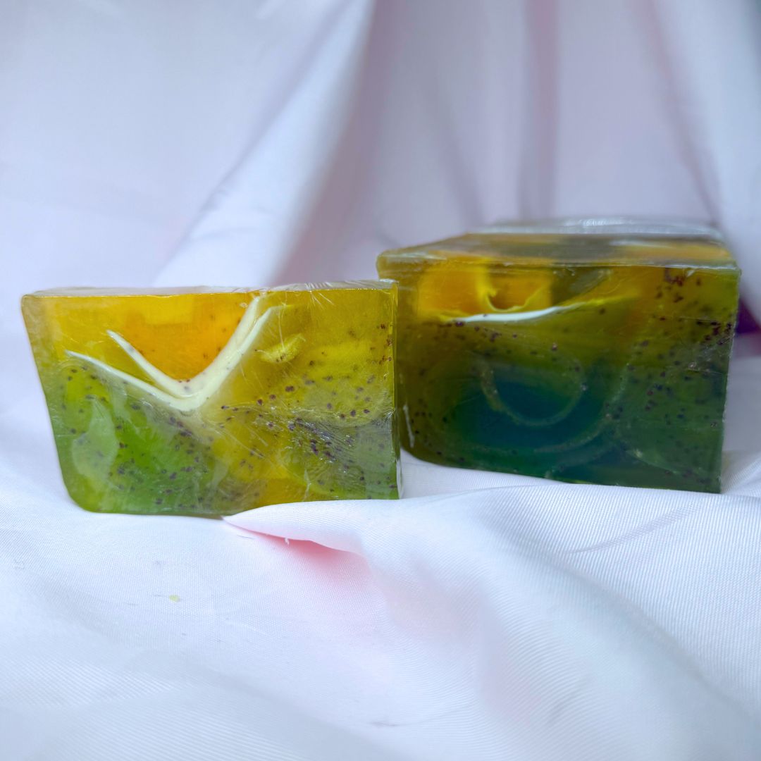 Bar of lemongrass cranberry soap with green and yellow gradient, black cranberry seeds, and white frills and larger soap loaf on a white fabric background