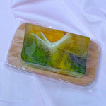 Bar of natural lemongrass cranberry soap with green and yellow gradient, black cranberry seeds, and white frills on a brown wooden soap dish and white fabric background