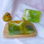Bar of lemongrass cranberry soap with green and yellow gradient, black cranberry seeds, and white frills on a wooden soap dish with beautiful Soap Rocks in Peridot and gold glitter rubber duck on a white background