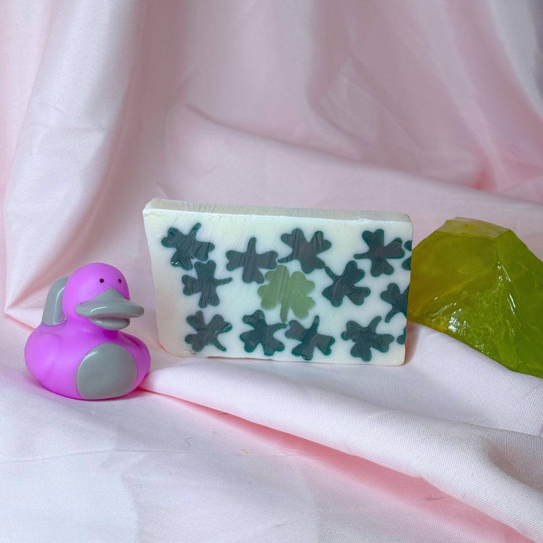 Luck of the irish saint patrick's St. Patrick's day bar soap with green clovers four leaf clovers, a purple platypus duck, and green Peridot Soap Rocks, and a pink fabric backdrop