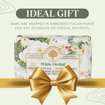 A gift set of White Orchid natural soap bars wrapped in decorative paper with a large gold ribbon and bow. The packaging features white orchid flowers and green leaves on a pale green background, with text reading 'IDEAL GIFT' at the top.