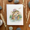 aubree sure art greeting card with watercolor illustration of dungeons and dragons grung frogfolk peaking out behind a lily pad lotus hat to look at a dragonfly. he holds a spear and wears a chalic of stone shards. blank inside with envelope.