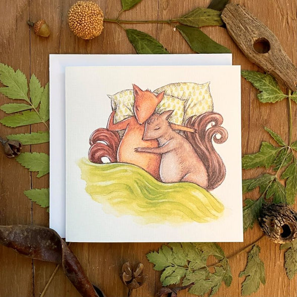 aubree sue art greeting card illustrated with watercolor design of two squirrels snuggling in bed with green blanket. great for anniversary or valentine's day. comes blank inside with envelope.